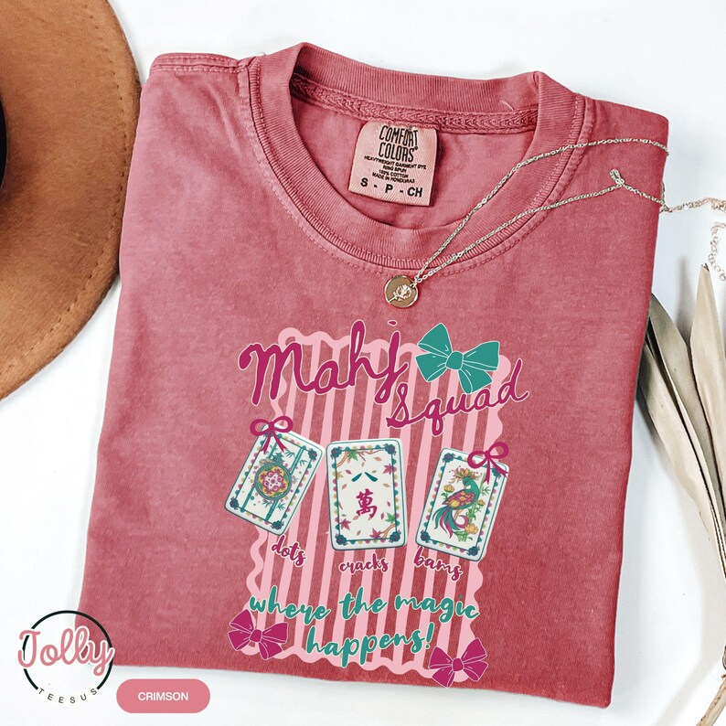 May include: Crimson-colored t-shirt with the text "Mahj Squad" and three illustrated Mahjong tiles. The shirt also features the phrase "where the magic happens" and decorative bows. A silver necklace with a pendant is draped across the shirt.