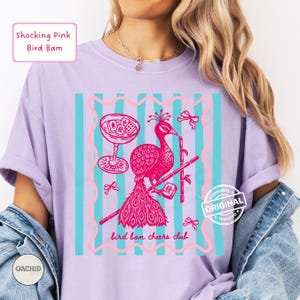 May include: A lilac-colored t-shirt with a graphic featuring a pink peacock, cocktail glass, and bamboo. The design includes the text "Shocking Pink Bird Bam" and "bird bam cheers club". The shirt has a relaxed fit.