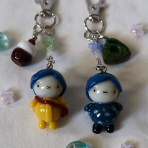May include: Two keychains featuring small, stylized figures with blue hair and white faces. One figure wears a yellow coat with a red strap, the other a dark blue star-patterned outfit. Both have silver lobster clasps and small decorative beads.