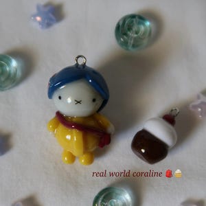 May include: A small figurine charm featuring a character with blue hair, a white face with an X-shaped mouth, and a yellow raincoat with a red strap, alongside a brown and white cupcake charm, with the text real world coraline and star beads.