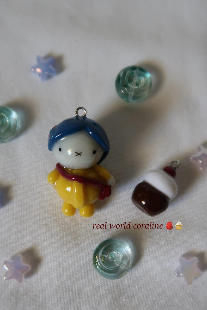 May include: A small figurine charm featuring a character with blue hair, a white face with an X-shaped mouth, and a yellow raincoat with a red strap, alongside a brown and white cupcake charm, with the text real world coraline and star beads.