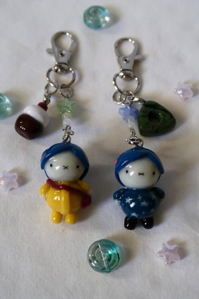 May include: Two keychains featuring small, stylized figures with blue hair and white faces. One figure wears a yellow coat with a red strap, the other a dark blue star-patterned outfit. Both have silver lobster clasps and small decorative beads.
