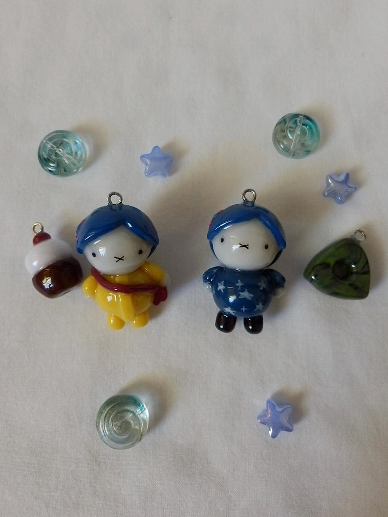 May include: Two glass character charms, one in a yellow outfit and one in a blue star-patterned outfit, accompanied by a brown and white cupcake charm, a green triangular charm, three light blue star beads, and two clear glass beads.