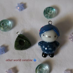 May include: A small figurine pendant with blue hair, a white face, and a dark blue star-patterned outfit, accompanied by a dark green triangular charm, several translucent light blue star beads, and clear glass beads, with the text other world coraline.