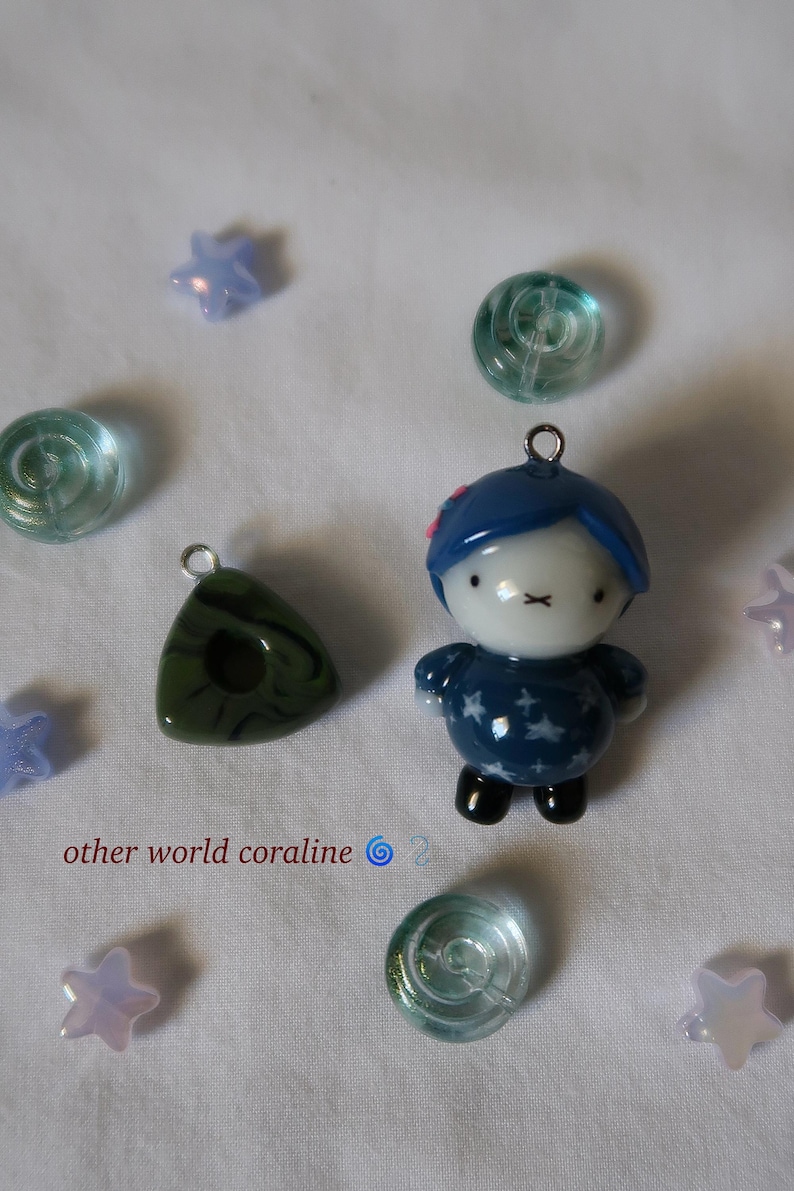 May include: A small figurine pendant with blue hair, a white face, and a dark blue star-patterned outfit, accompanied by a dark green triangular charm, several translucent light blue star beads, and clear glass beads, with the text other world coraline.