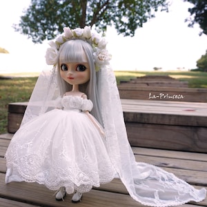 Può includere: Una bambola di porcellana bianca che indossa un abito da sposa in pizzo bianco e un velo. La bambola ha lunghi capelli argentati e una corona di fiori. La bambola è in piedi su una superficie di legno.