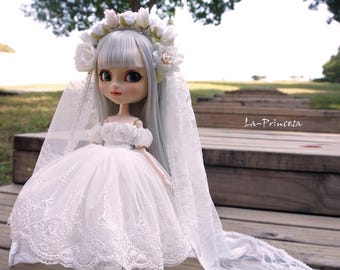 Peut inclure: Une poupée en porcelaine blanche portant une robe de mariée en dentelle blanche et un voile. La poupée a de longs cheveux argentés et une couronne de fleurs. La poupée est debout sur une surface en bois.