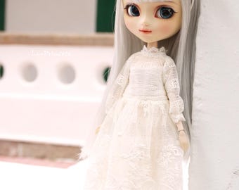 Peut inclure: Une poupée aux longs cheveux argentés et aux yeux bleus, vêtue d'une robe en dentelle blanche et d'un couvre-chef assorti. La poupée est debout à côté d'un mur blanc. Les chaussures de la poupée sont blanches avec des semelles noires.