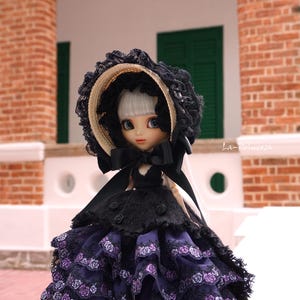 May include: A doll in a black dress with a layered purple skirt featuring floral lace. The doll has a black hat with lace trim and a large bow. The doll has pale skin, blue eyes, and white hair. The text "La-Princesa" is visible.