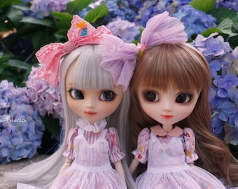 Peut inclure: Deux poupées aux cheveux longs portant des robes et des bandeaux roses et blancs. Les poupées sont assises devant un buisson d'hortensias bleus.