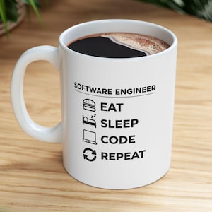 May include: White ceramic mug filled with black coffee, featuring the text "SOFTWARE ENGINEER" and the words "EAT SLEEP CODE REPEAT" with icons. The mug has a white handle and is on a wooden surface.