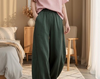 Puede incluir: Una persona viste una camiseta rosa claro de manga corta y pantalones verdes oscuros de pernera ancha. Los pantalones tienen una cintura elástica. Zapatillas blancas completan el atuendo. El escenario es un dormitorio con una paleta de colores neutros.