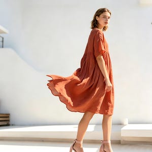 Puede incluir: Vestido corto de color óxido, con mangas cortas y falda fluida. El vestido tiene un corte holgado y cómodo y se combina con sandalias de tacón rosa claro. La modelo está de pie frente a una pared blanca, con la tela del vestido ondeando al viento.