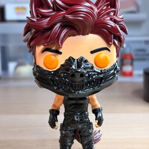 Può includere: Una statuetta da collezione in vinile stilizzata che raffigura un personaggio con capelli rosso scuro a punta, orecchie da lupo e occhi arancioni brillanti. Il personaggio indossa una maschera da lupo nera, un top senza maniche e pantaloni neri.