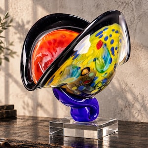 Könnte beinhalten: Eine dekorative Glasskulptur mit einer breiten, geschwungenen Schalenform und einem schwarzen Rand. Das Innere zeigt einen leuchtend orangefarbenen Bereich und eine gelbe Oberfläche mit blauen, roten und grünen Punkten. Sie ruht auf einem blauen Glassockel.