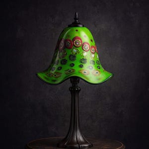 Könnte beinhalten: Eine grüne Glaslampe mit floralem Design auf einem dunkelbraunen Tisch. Die Lampe hat einen ausgestellten Schirm mit roten und gelben Blumenmustern. Der Sockel ist dunkelbraun und verziert. Der Tisch ist rund und aus Holz.