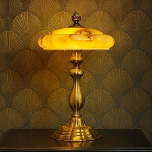 May include: A vintage-style table lamp with a golden base and a yellow, rounded shade. The lamp has an ornate, detailed design and sits on a dark table. The background features a patterned wallpaper.