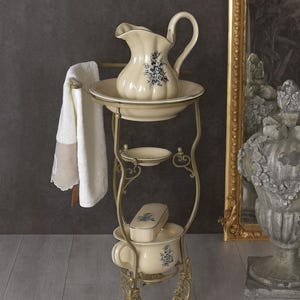Könnte beinhalten: Ein cremefarbener Waschständer im Vintage-Stil mit passendem Krug, Waschbecken und Seifenschale, verziert mit blauen Blumenmustern. Ein weißes Handtuch hängt an der Seite. Der Ständer ist aus verziertem goldfarbenem Metall.