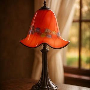 Könnte beinhalten: Eine dekorative Tischlampe mit einem leuchtend orangefarbenen Glasschirm. Der Schirm hat farbenfrohe, abstrakte Designs und einen gewellten Rand. Die Lampe hat einen dunklen, verzierten Fuß und steht auf einem Holztisch. Die Lampe ist eine dekorative Leuchte.