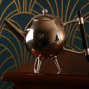 Könnte beinhalten: Eine polierte silberne Teekanne mit rundem Körper und Ausguss. Die Teekanne hat einen schwarzen Griff und steht auf drei kleinen Beinen. Der Hintergrund zeigt ein teal-goldenes Art-Deco-Muster. Es ist ein Küchenartikel.