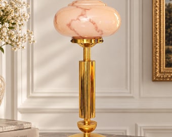 Puede incluir: Una lámpara de mesa con base de metal dorado pulido, vástago cilíndrico y pantalla de cristal redondeada en color rosa pálido con vetas marmoladas. La lámpara cuenta con un pequeño remate dorado en la parte superior y una base circular.