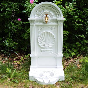 May include: A white outdoor water fountain with ornate detailing, including a shell design and fluted columns. The fountain has a brass faucet and is set against a backdrop of lush greenery and pink flowers.