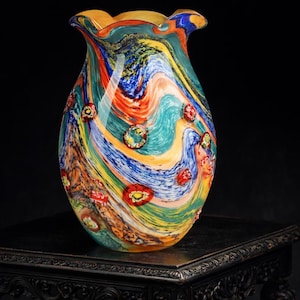Könnte beinhalten: Eine farbenfrohe, mundgeblasene Glasvase mit gewelltem Rand. Die Vase zeigt wirbelnde Muster aus Blau, Orange, Gelb und Grün mit kleinen, roten floralen Akzenten. Die dekorative Vase ist ein einzigartiges Kunstwerk.