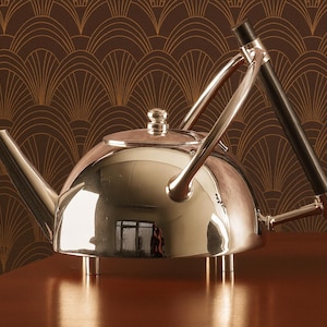 Könnte beinhalten: Eine glänzende Chrom-Teekanne mit schwarzem Griff und gebogenem Ausguss. Die Teekanne steht auf einer braunen Oberfläche, die das Licht reflektiert. Der Hintergrund zeigt ein braunes und goldenes Art-Deco-Muster.