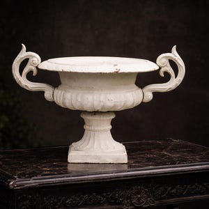 May include: A white, ornate urn with two curved handles, set on a dark, patterned table. The urn has a wide, flat top and a pedestal base. The background is a dark, blurred backdrop.