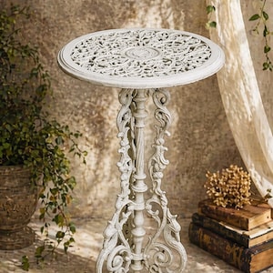 May include: A white, ornate cast iron side table with a round top featuring a detailed, openwork design. The table has a decorative, scrolled base and a central column. The table is approximately 71 cm tall.