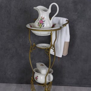 May include: A vintage white porcelain washbasin set with floral accents, including a pitcher, bowl, soap dish, and chamber pot. The set is displayed on a decorative brass stand with a towel rack. The stand is approximately 48 inches tall.