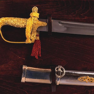 Op de afbeelding: Een decoratief zwaard met een goudkleurig handvat en accenten, een zilveren lemmet en een rode kwast. Het zwaard wordt getoond op een donkerhouten oppervlak, met de schede eronder.