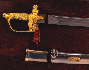 May include: A decorative sword with a gold handle and accents, a silver blade, and a red tassel. The sword is displayed on a dark wood surface, with the scabbard visible below.