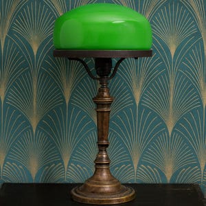 May include: An antique desk lamp with a green glass shade. The lamp has a bronze-coloured base and a dark brown metal support for the shade. The lamp is on a black table against a teal and gold patterned wallpaper.