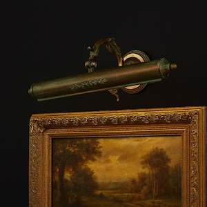 May include: An antique bronze picture light, mounted above a framed painting. The light has a cylindrical shade and decorative details. The painting is in a gold ornate frame. The light is designed to illuminate artwork.