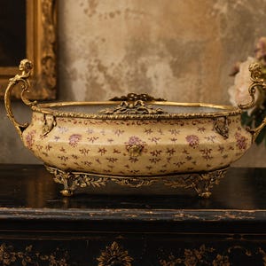 Puede incluir: Un cuenco decorativo ovalado de estilo antiguo con un estampado floral crema. El cuenco tiene asas doradas ornamentadas y una base decorativa. El cuenco está sobre una superficie negra.