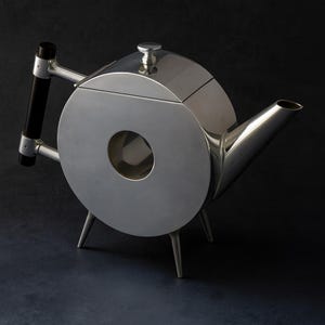 Op de afbeelding: Een moderne, zilverkleurige theepot met een cirkelvormig ontwerp en een zwarte handgreep. De theepot heeft een tuit, een deksel met een kleine knop en drie slanke poten. De unieke vorm en gepolijste afwerking geven het een eigentijdse uitstraling.