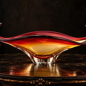 Peut inclure: Un bol décoratif en verre présentant une forme allongée en bateau avec des extrémités recourbées vers le haut. Le verre passe d'une teinte rouge profond sur le bord supérieur à une base ambrée translucide et claire, créant un dégradé.