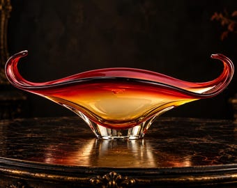 May include: A decorative glass bowl featuring an elongated, boat-like shape with upward-curving ends. The glass transitions from a deep red hue at the top rim to a warm, translucent amber and clear base, creating a smooth gradient effect.