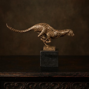 May include: A bronze sculpture of a cheetah in mid-stride, with a spotted pattern. The sculpture is mounted on a black square base, and the cheetah's form is elongated and dynamic. The background is a dark, textured brown.