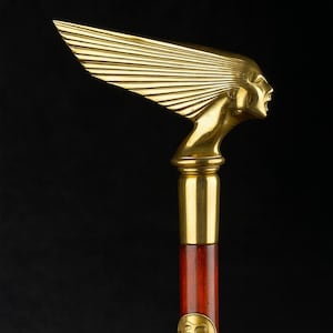 Puede incluir: Bastón dorado con un diseño estilizado de cabeza y alas. La cabeza está en la parte superior, con un mango de madera y un emblema circular en la parte inferior. El bastón tiene un fondo oscuro.