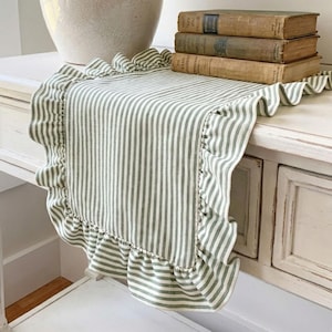 Peut inclure: Une table blanche avec un chemin de table à rayures vertes et blanches avec des bords à volants. Une lampe beige et trois livres anciens sont sur la table. La table a un aspect vieilli et vintage.