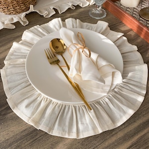 May include: A dining table setting with a white plate, gold-coloured cutlery, and a folded white napkin tied with twine. The plate sits on a cream placemat with a frilled edge. The setting is on a wooden table.