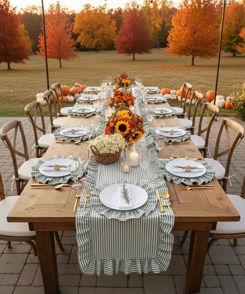 Peut inclure: Une longue table en bois dress&eacute;e pour un repas en plein air &agrave; l'automne. La table est orn&eacute;e d'assiettes blanches, de couverts dor&eacute;s et de sets de table &agrave; rayures vertes et blanches. Des centres de table avec des tournesols et des fleurs d'automne.