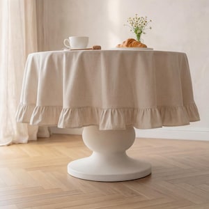 May include: A round, white pedestal table with a beige linen tablecloth. The tablecloth has a ruffled edge. A white teacup, honey dipper, croissant, and small vase of flowers are on the table.