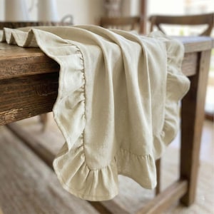May include: A beige linen table runner with ruffled edges draped over a wooden table. The runner has a soft, flowing texture and adds a touch of elegance to the table setting. The neutral color complements the rustic wood.