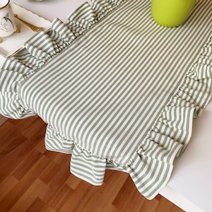 May include: A green and white striped table runner with frilled edges. The runner is made of fabric and features a classic stripe pattern. The frilled edges add a decorative touch to the table setting. The runner is placed on a white surface.