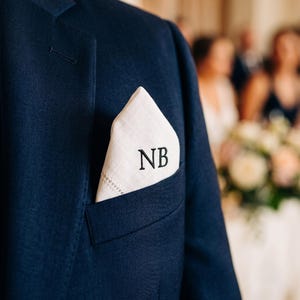 May include: A white linen pocket square with a decorative hemstitch border and the embroidered black initials NB, tucked into the breast pocket of a dark navy blue suit jacket. The fabric has a visible woven texture and a crisp, folded point.