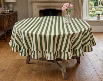 May include: A round tablecloth featuring vertical dark green and white stripes with a ruffled edge. The fabric drapes over a wooden table, and a clear glass vase containing three pink flowers sits on the center of the tabletop surface.