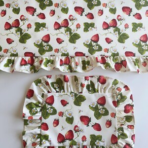 May include: A white fabric table runner and placemat set. The set features a repeating pattern of red strawberries, green leaves, and white flowers. Both pieces have a ruffled edge.